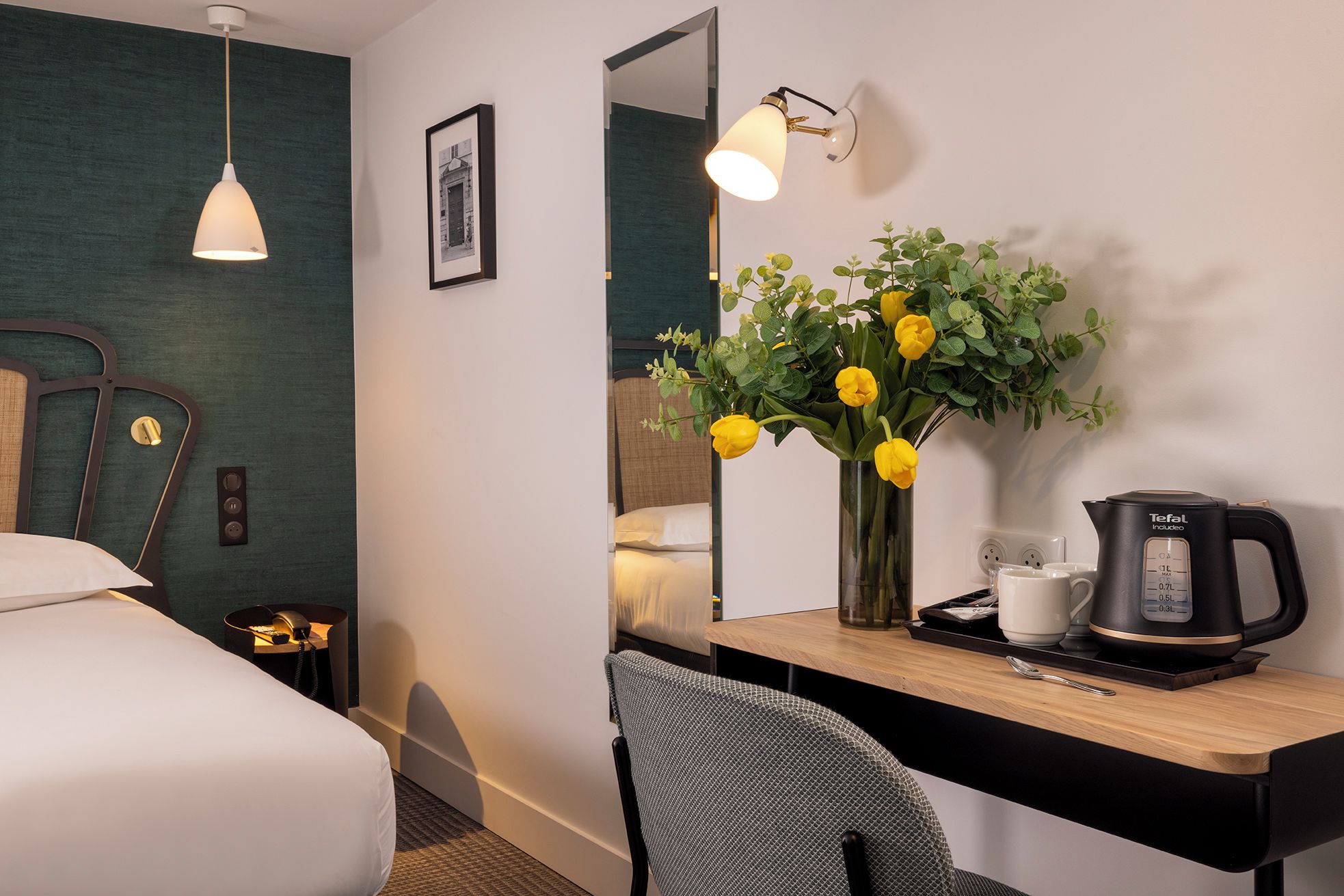 A hotel room with a bed, a desk with a coffee maker, a chair, a mirror, and a vase with flowers.