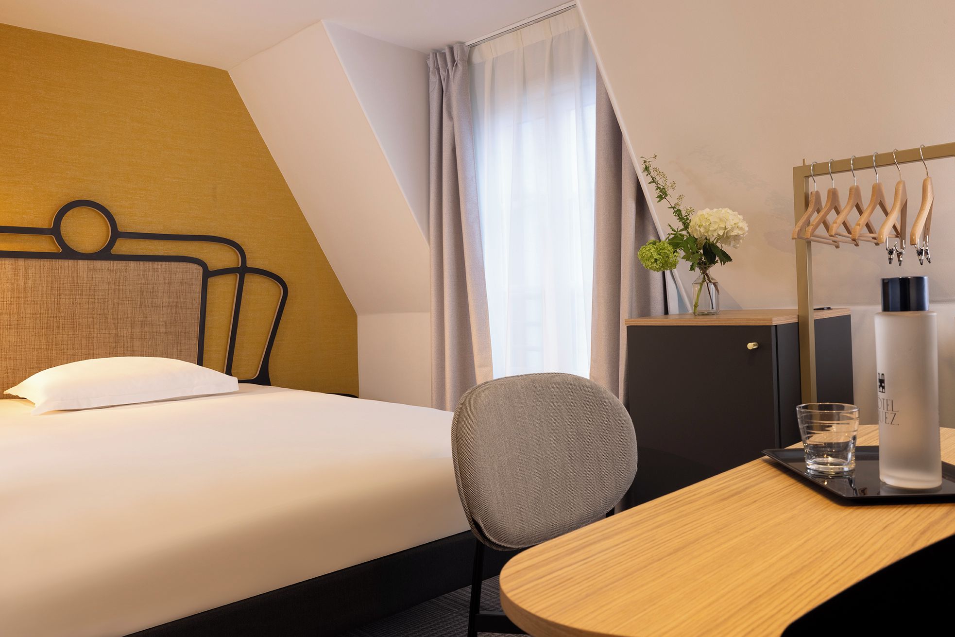 Habitación de hotel con cama, escritorio, silla, armario, espejo y ventana con cortinas.