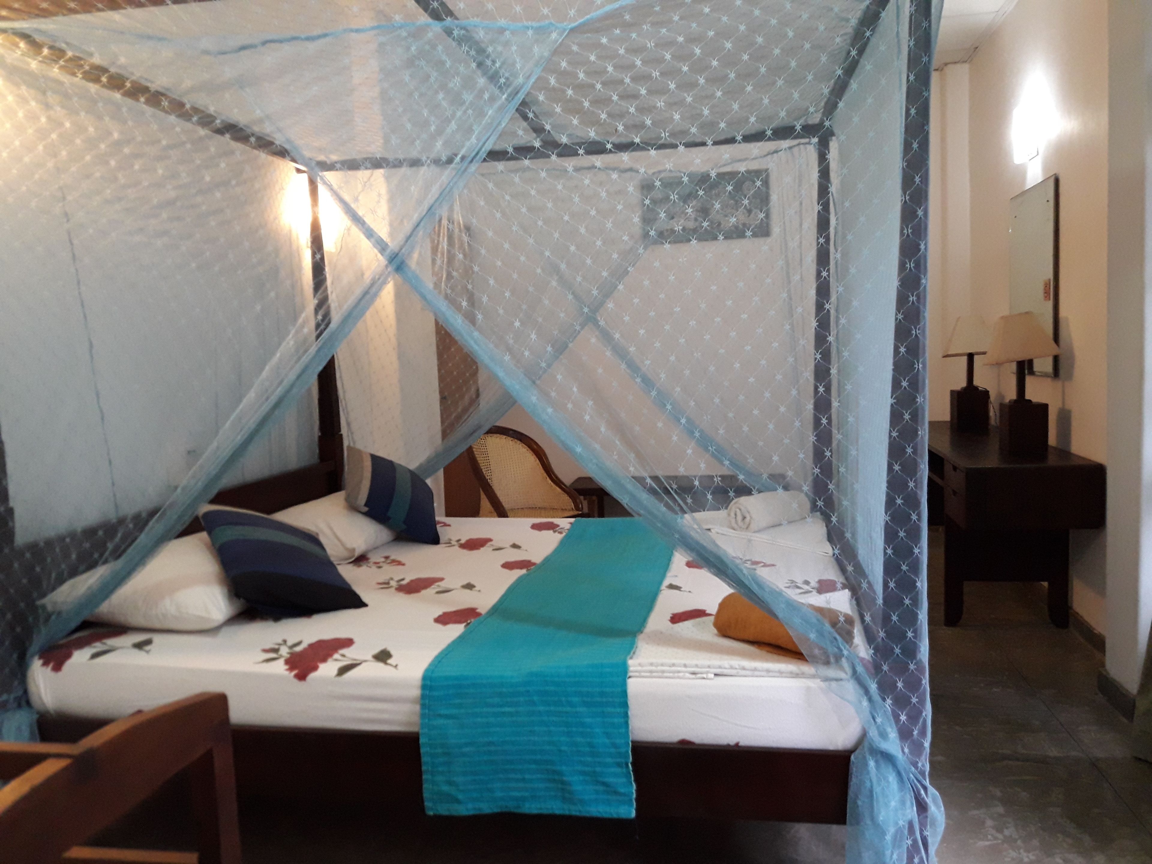 A bed with a mosquito net, a wooden chair, a nightstand with a lamp, and a wall-mounted mirror.