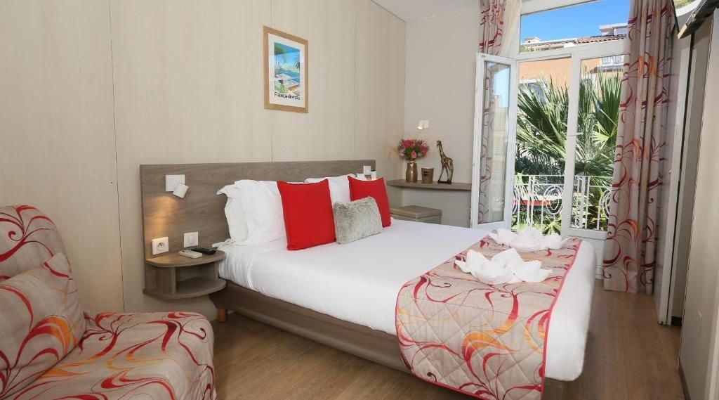A hotel room with a bed, bedside tables, a nightstand, a window with curtains, and a view of palm trees.