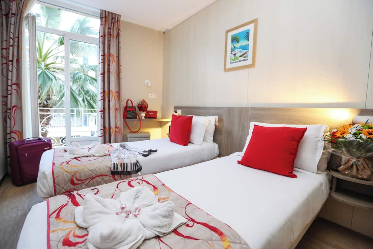 Ein Hotelzimmer mit zwei Betten, einem Fenster mit Vorhängen, einem Bild an der Wand und einem Nachttisch mit Blumen.