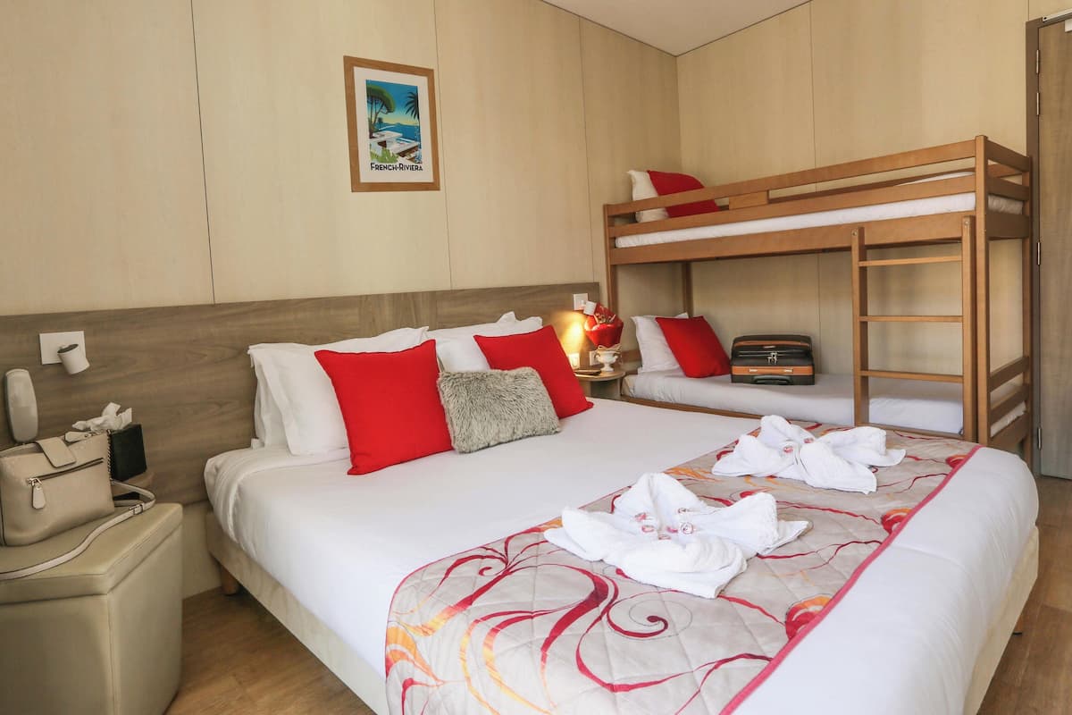 Ein Hotelzimmer mit einem Etagenbett, einem Nachttisch mit Lampe, einem gerahmten Bild an der Wand und einem ordentlich bezogenen Bett mit blumigem Muster.