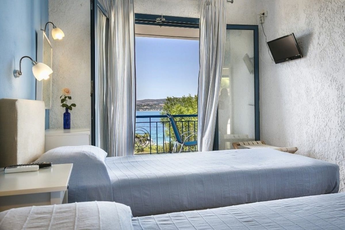 A hotel room with a bed, a desk, a lamp, a TV, and a balcony with a view of the sea.