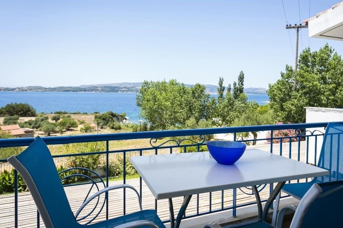 A balcony with a table and chairs, overlooking a body of water and greenery.