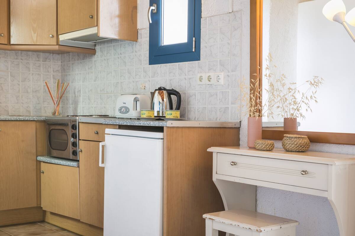A compact kitchen with wooden cabinets, a white refrigerator, a microwave, and a toaster. There is a small table with baskets and a potted plant.