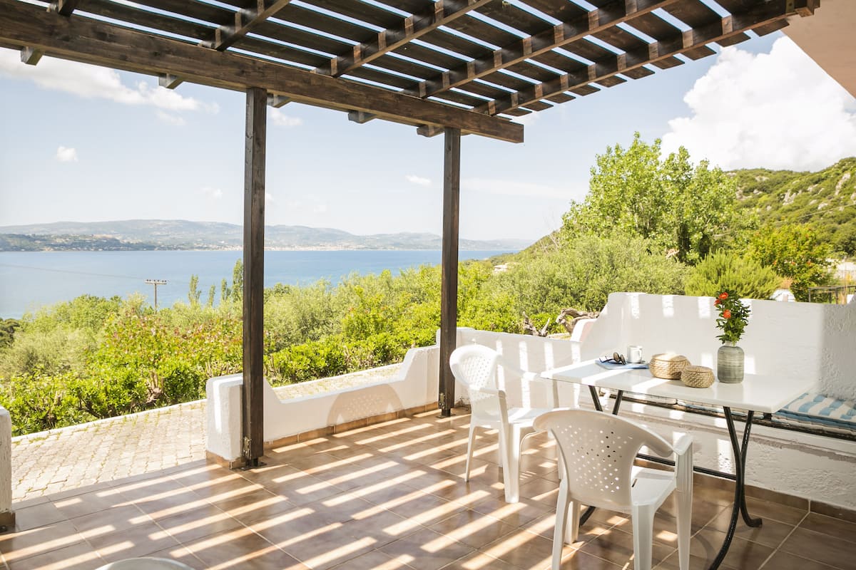 A covered patio with a table and chairs, overlooking a scenic view of the sea and hills.