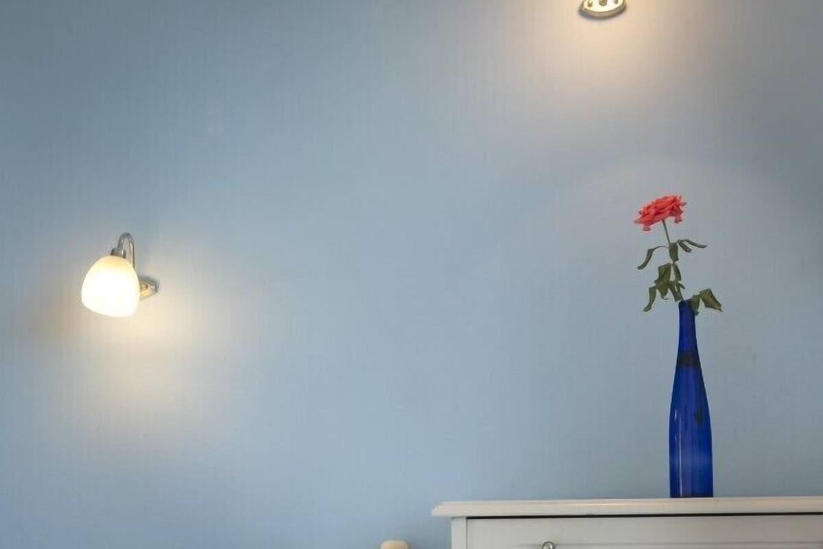 A bedroom with a bed, a nightstand, a small table, and a vase with a flower.