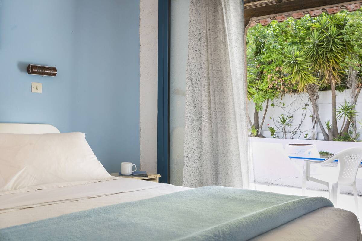 A bedroom with a bed, a nightstand, a cup, and a view of a balcony with plants and a chair.