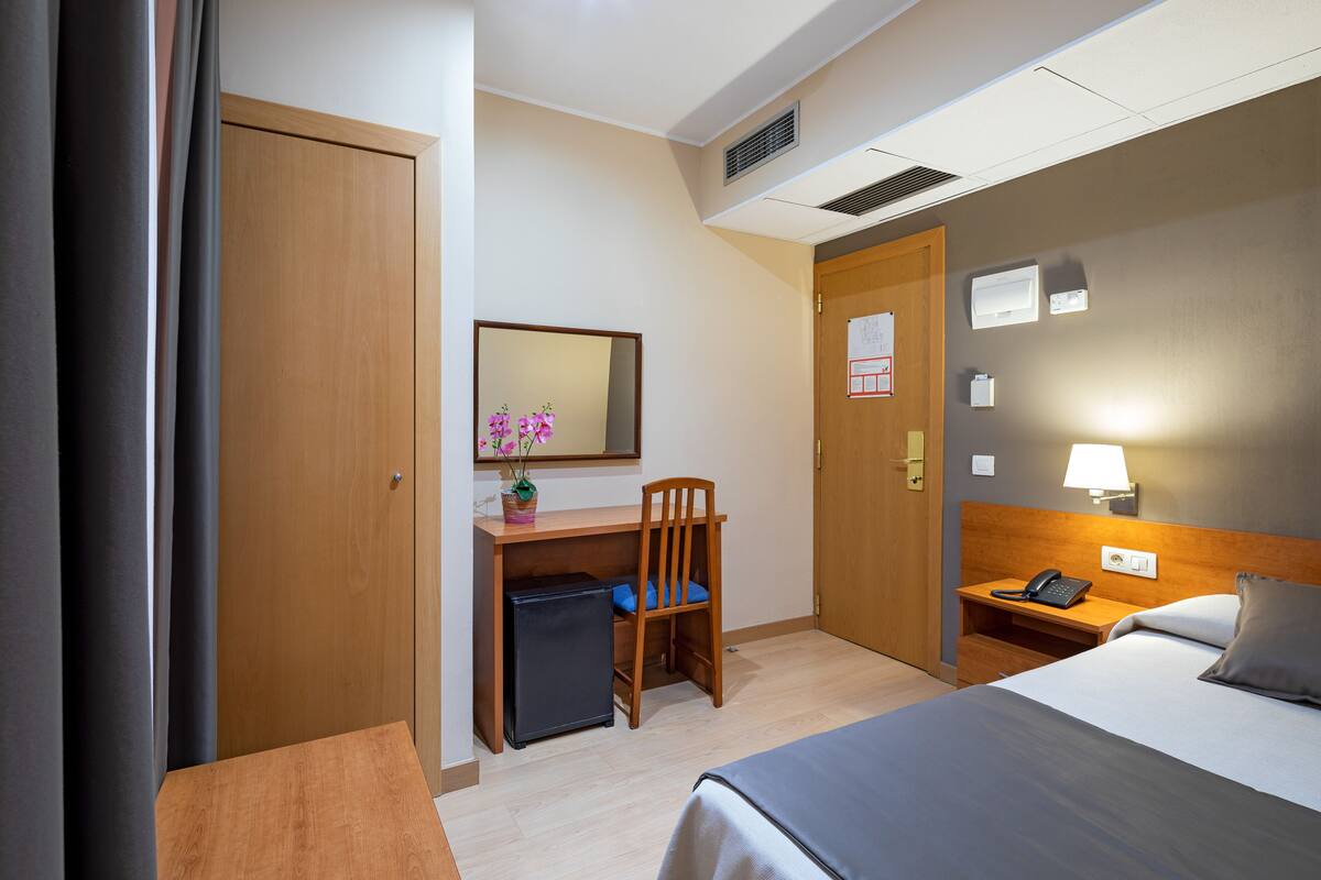 A hotel room with a bed, desk, chair, and a small refrigerator.
