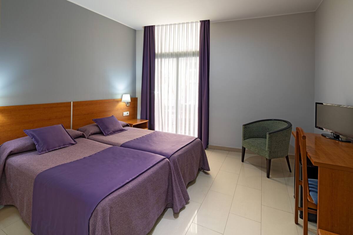 A hotel room with two beds, a desk, a chair, a TV, and a window with curtains.