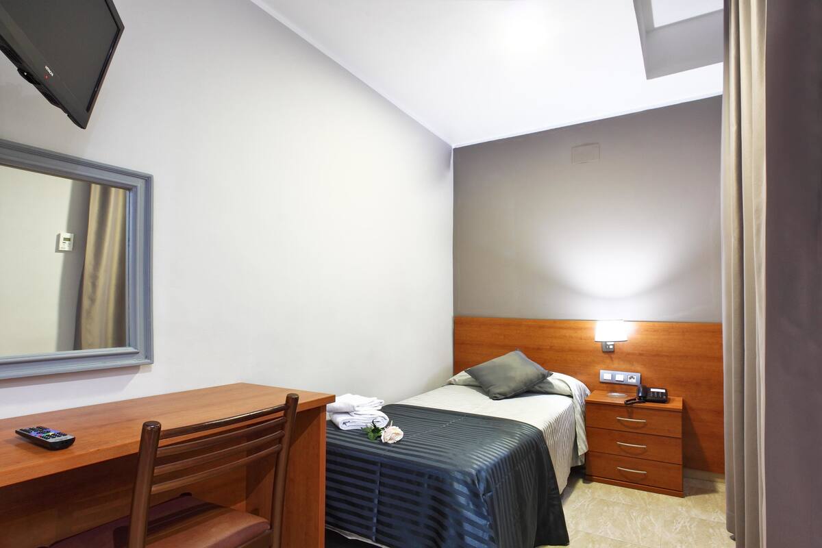 Un dormitorio compacto con un escritorio de madera, una cama con cabecera, una mesita de noche y un espejo.