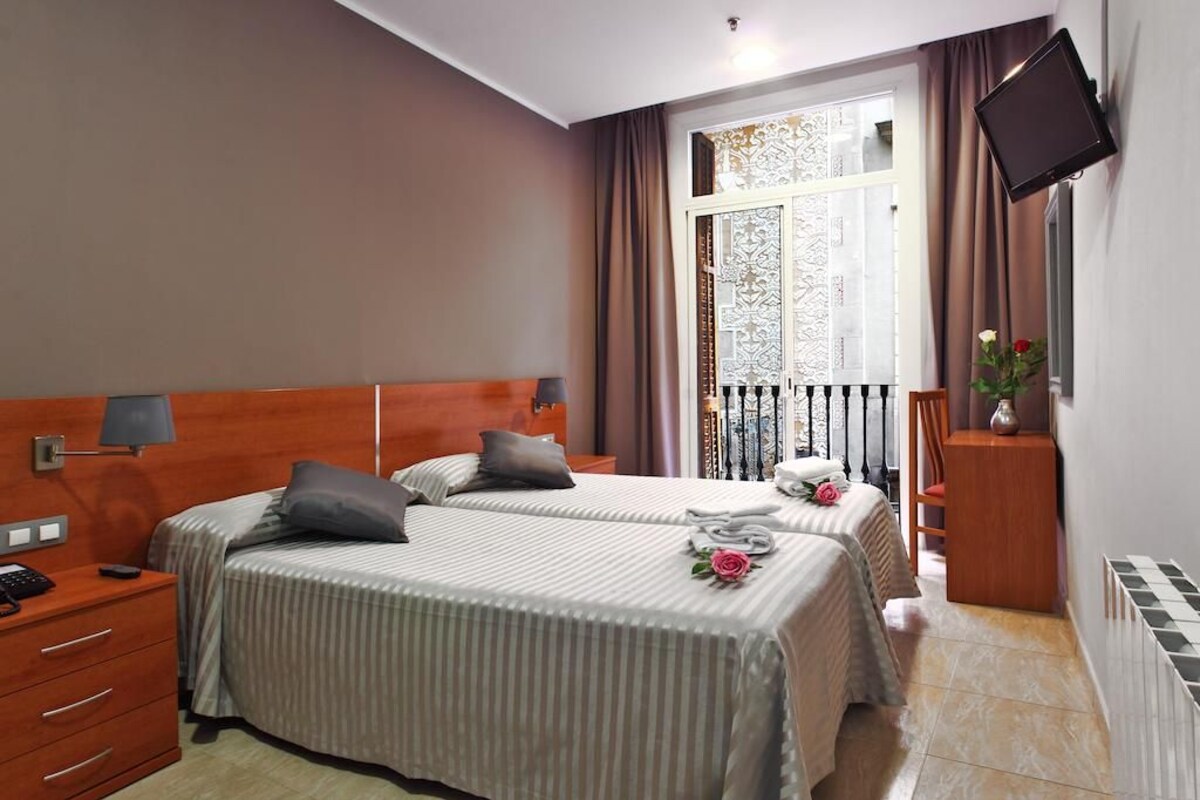 Quarto de hotel com duas camas, televisão na parede, janela com cortinas e uma mesinha com um vaso.