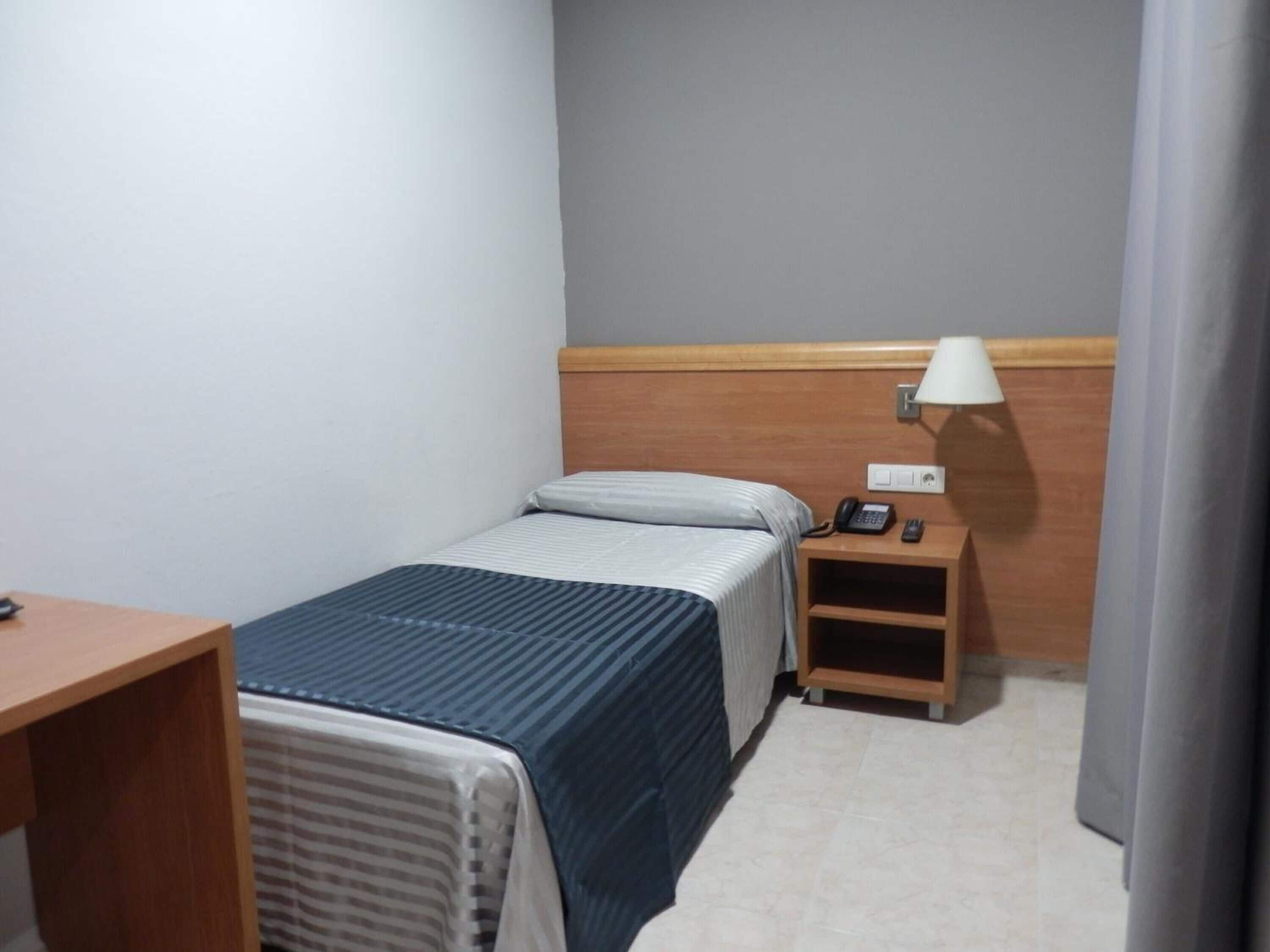 A small, neatly arranged hotel room with a single bed, a bedside table with a lamp, a phone, and a small wooden nightstand.