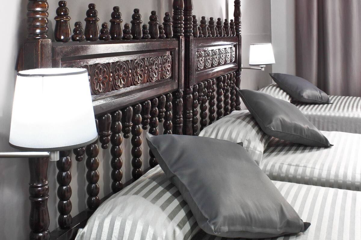 Una habitación de hotel con cabecera de madera, dos camas con ropa de cama a rayas, una mesita de noche con lámpara y una ventana con cortinas.