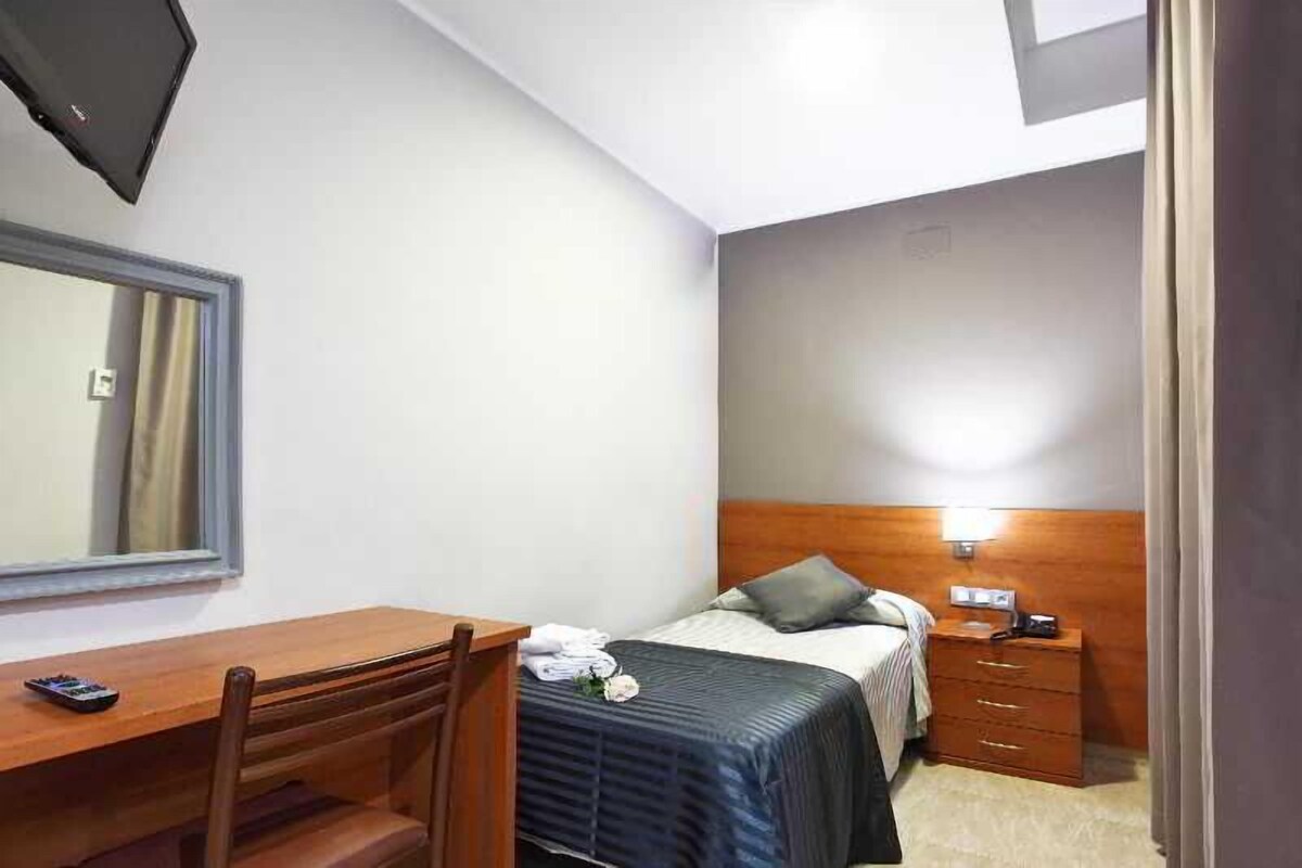 Quarto de hotel pequeno e moderno com uma cama de solteiro, uma mesa de cabeceira de madeira, uma escrivaninha com cadeira e um espelho.
