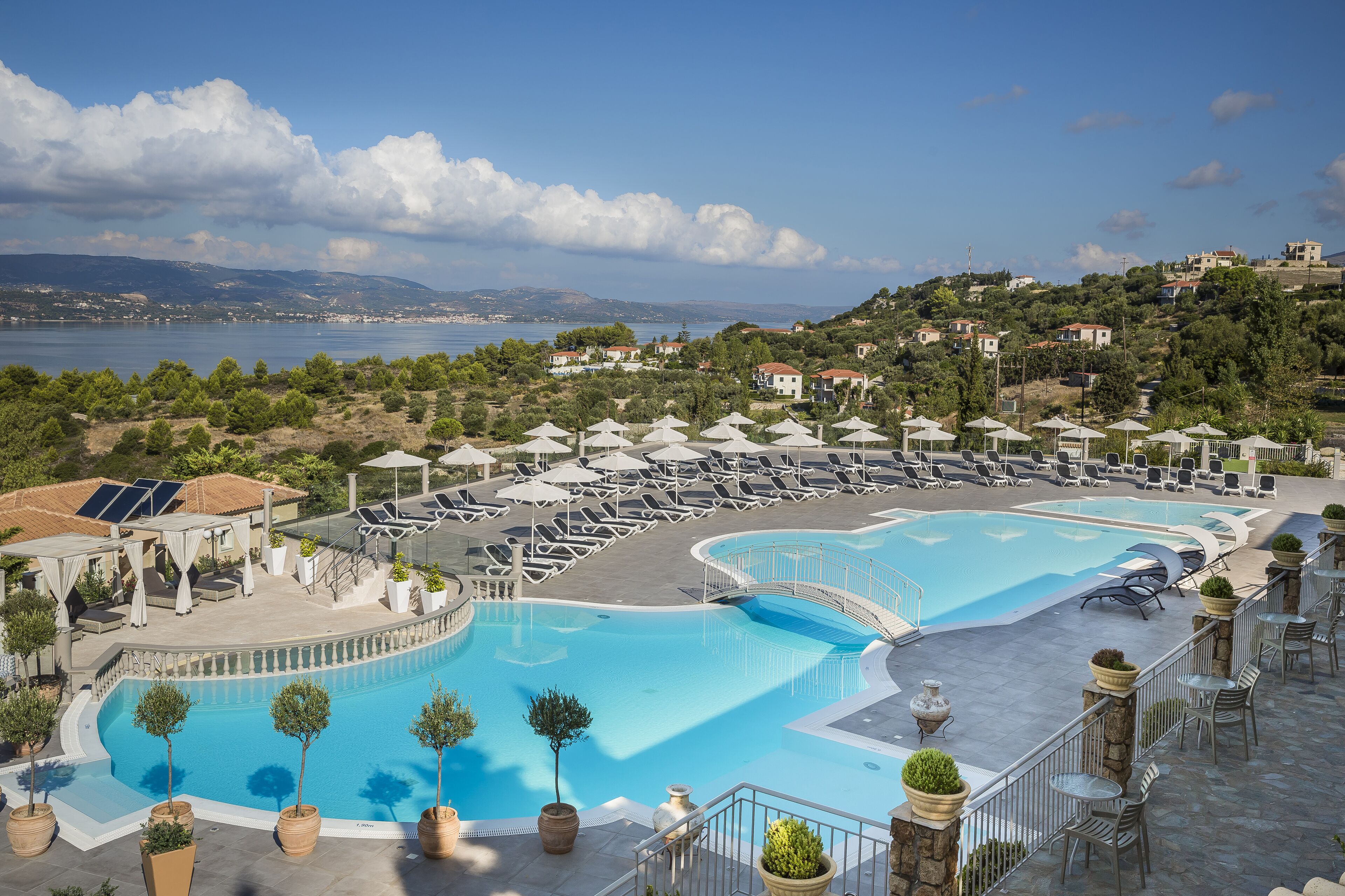 Ein Resort mit mehreren Schwimmbecken, Liegestühlen und Sonnenschirmen, umgeben von Hügeln und Blick auf das Meer.