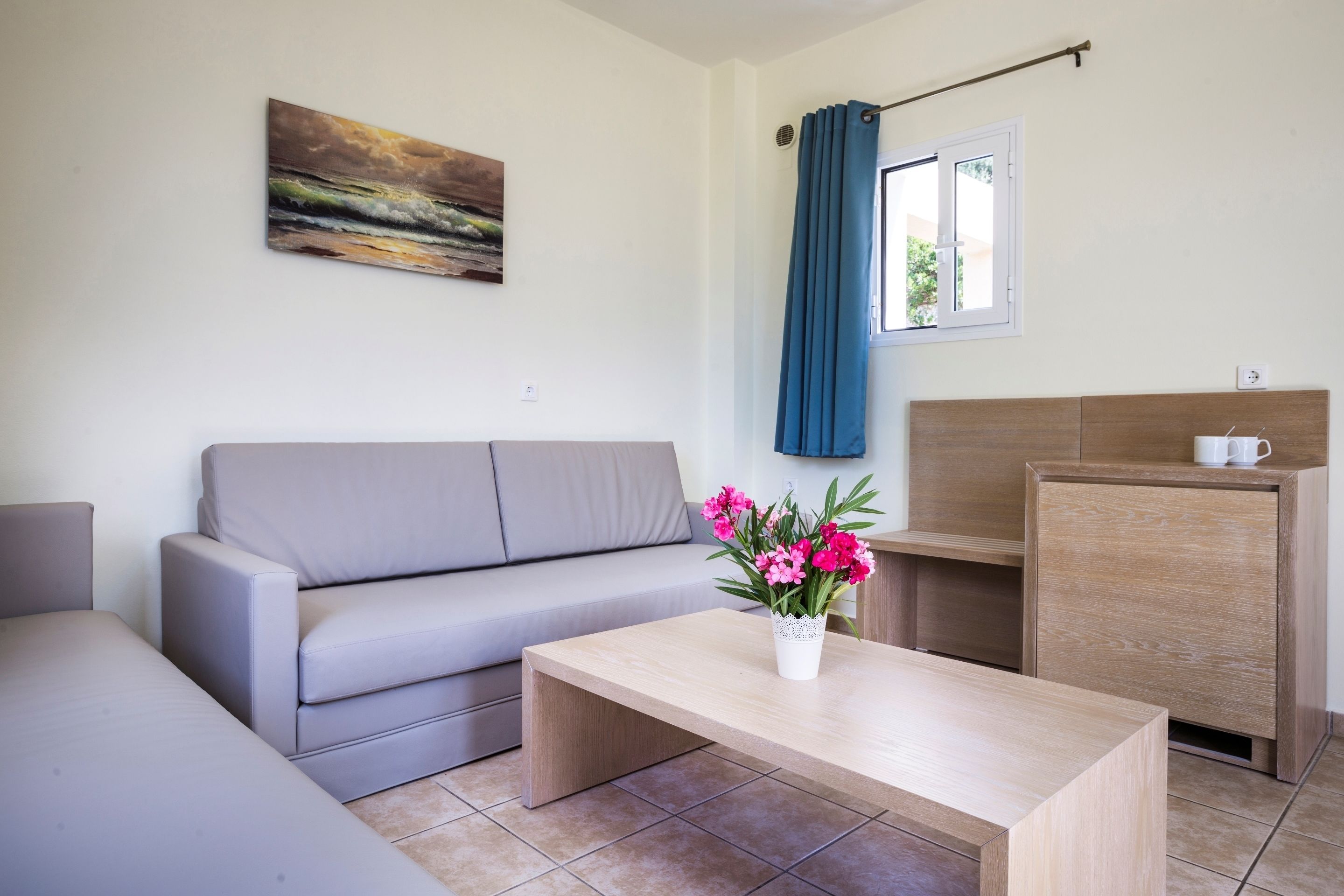 Un salon moderne avec un canapé gris, une table basse en bois ornée d’un vase de fleurs, un meuble en bois et un tableau accroché au mur.