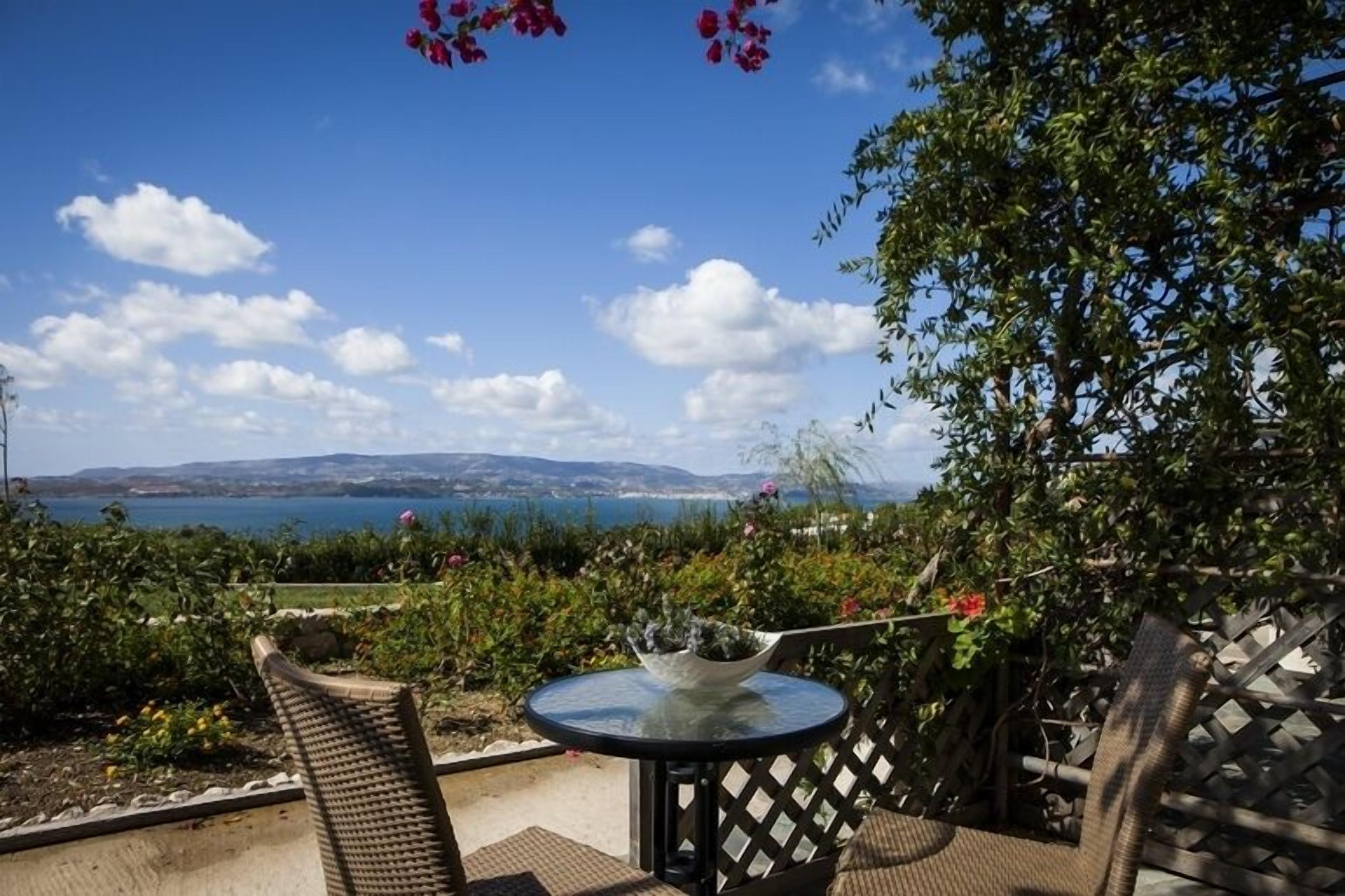 Une terrasse avec une table ronde et des chaises, offrant une vue panoramique sur un plan d’eau et des collines lointaines.
