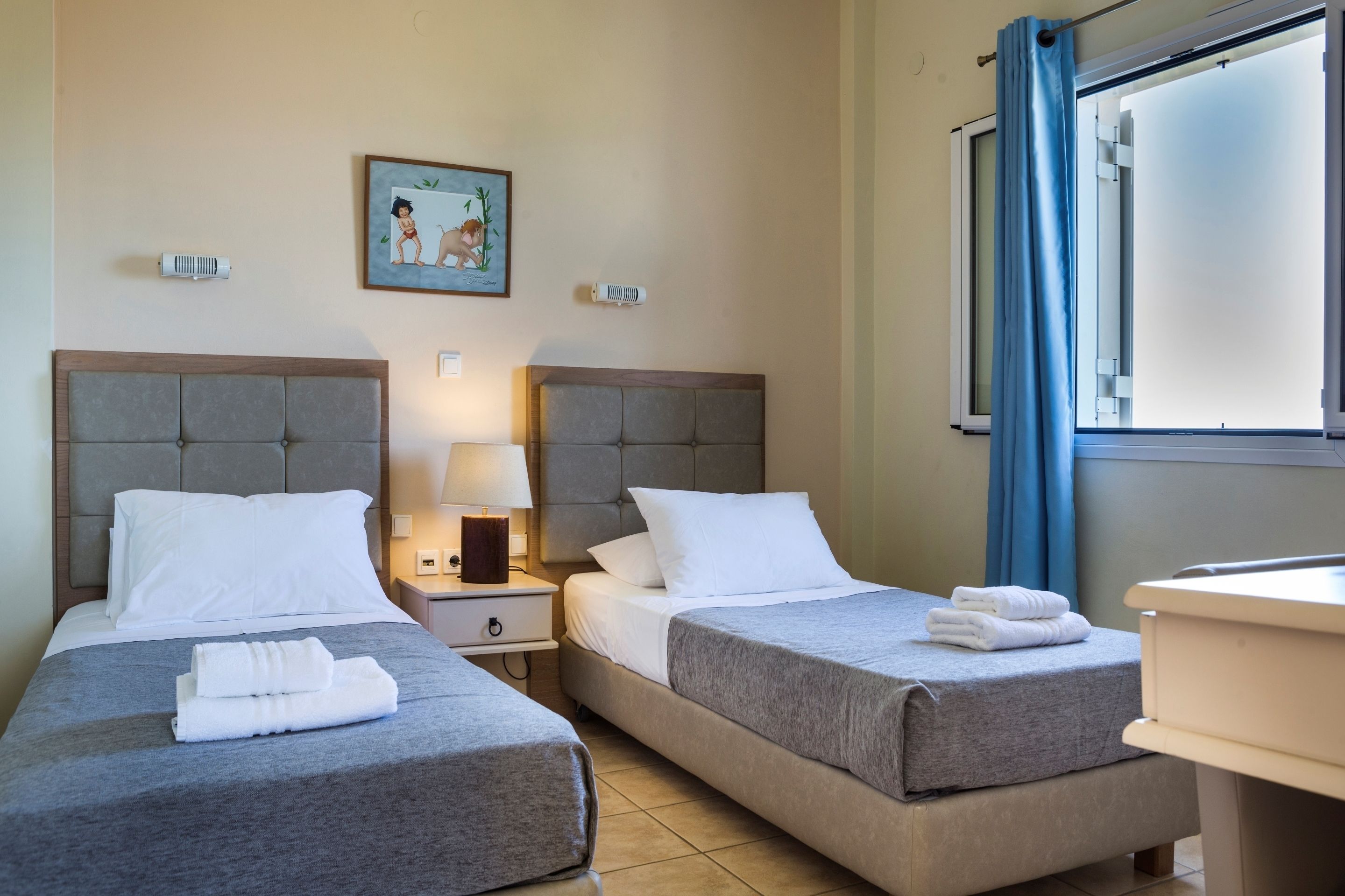 A hotel room with two beds, a nightstand with a lamp, a picture on the wall, and a window with curtains.