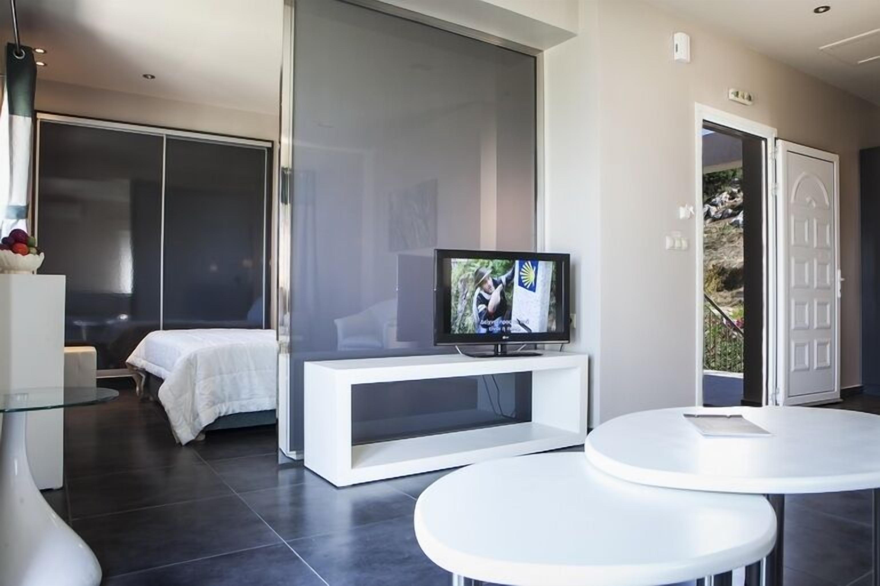 A modern hotel room with a bed, a TV on a white stand, a glass table, and a sliding door leading to a balcony.