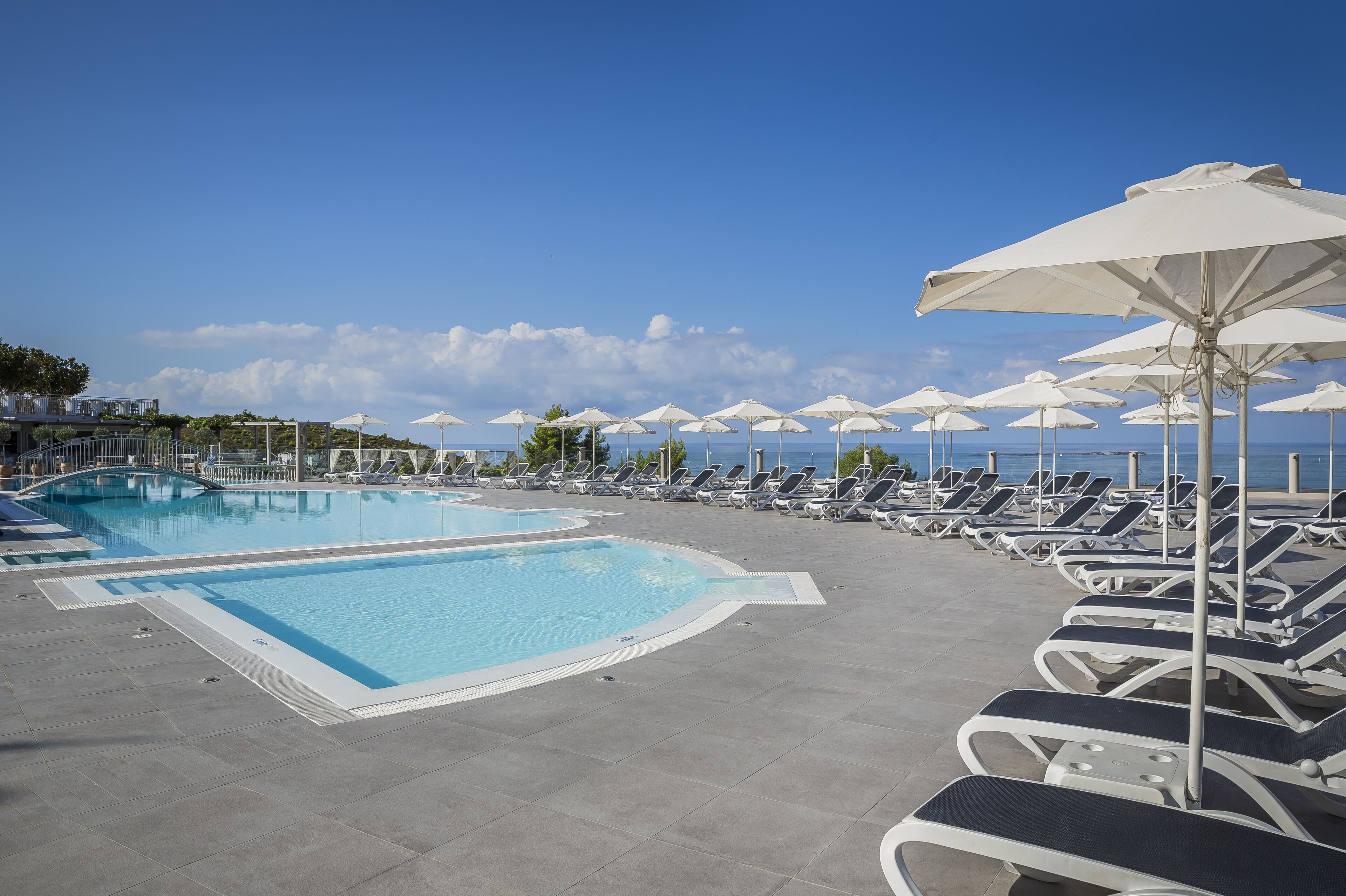 Un espace piscine avec des chaises longues et des parasols, un ciel dégagé et une vue sur la mer.