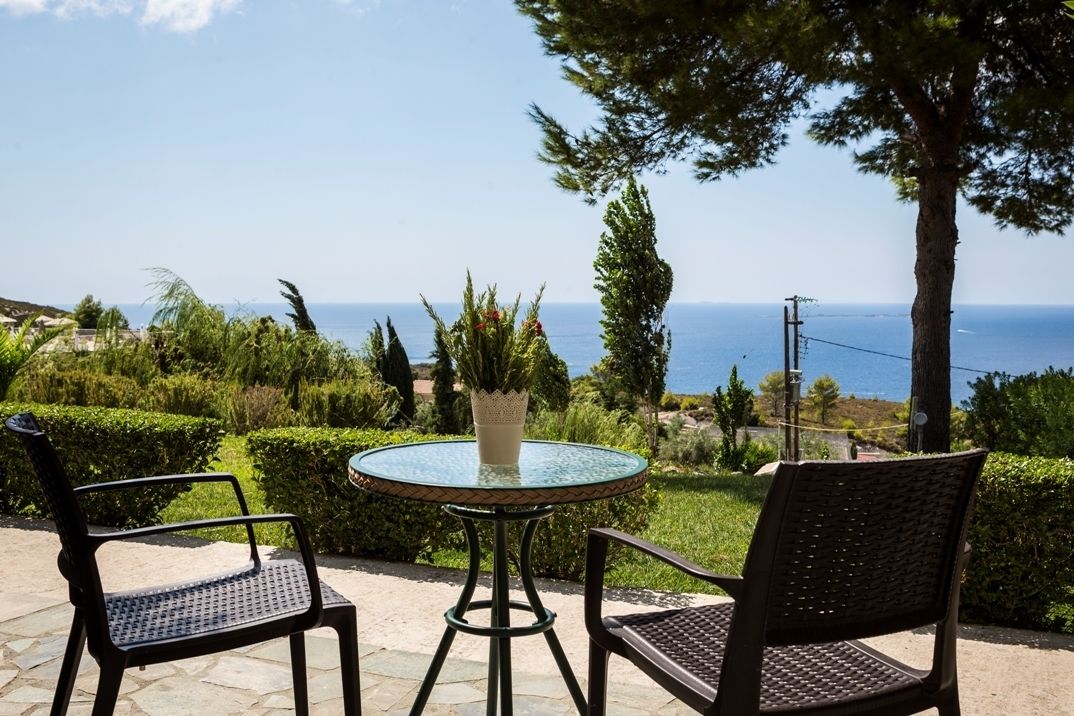 Une table ronde en verre, accompagnée de deux chaises, d’une plante en pot et offrant une vue sur la mer.