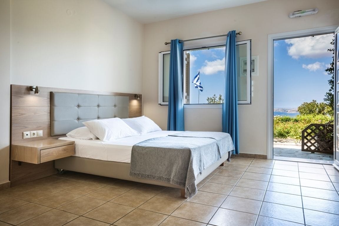Quarto com cama, mesa de cabeceira, janela com cortinas e vista para o exterior.