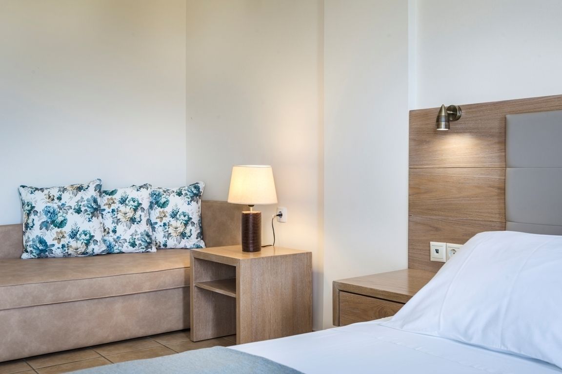 Hotelkamer met een bed, nachtkastje met lamp, houten hoofdeinde en een bank met bloemenkussens.
