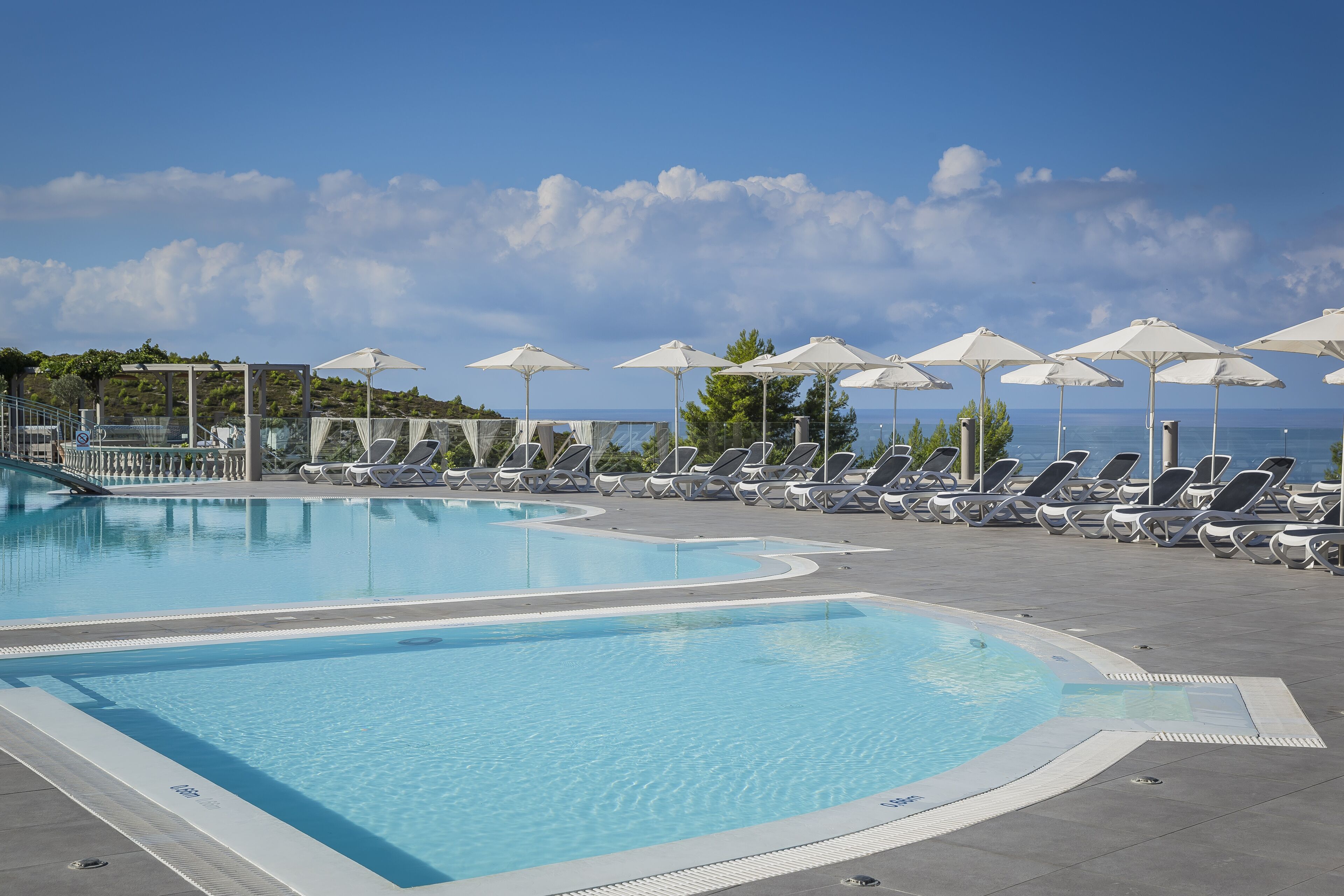 Una piscina con sillas de descanso y sombrillas, rodeada por una terraza pavimentada y vistas al mar.
