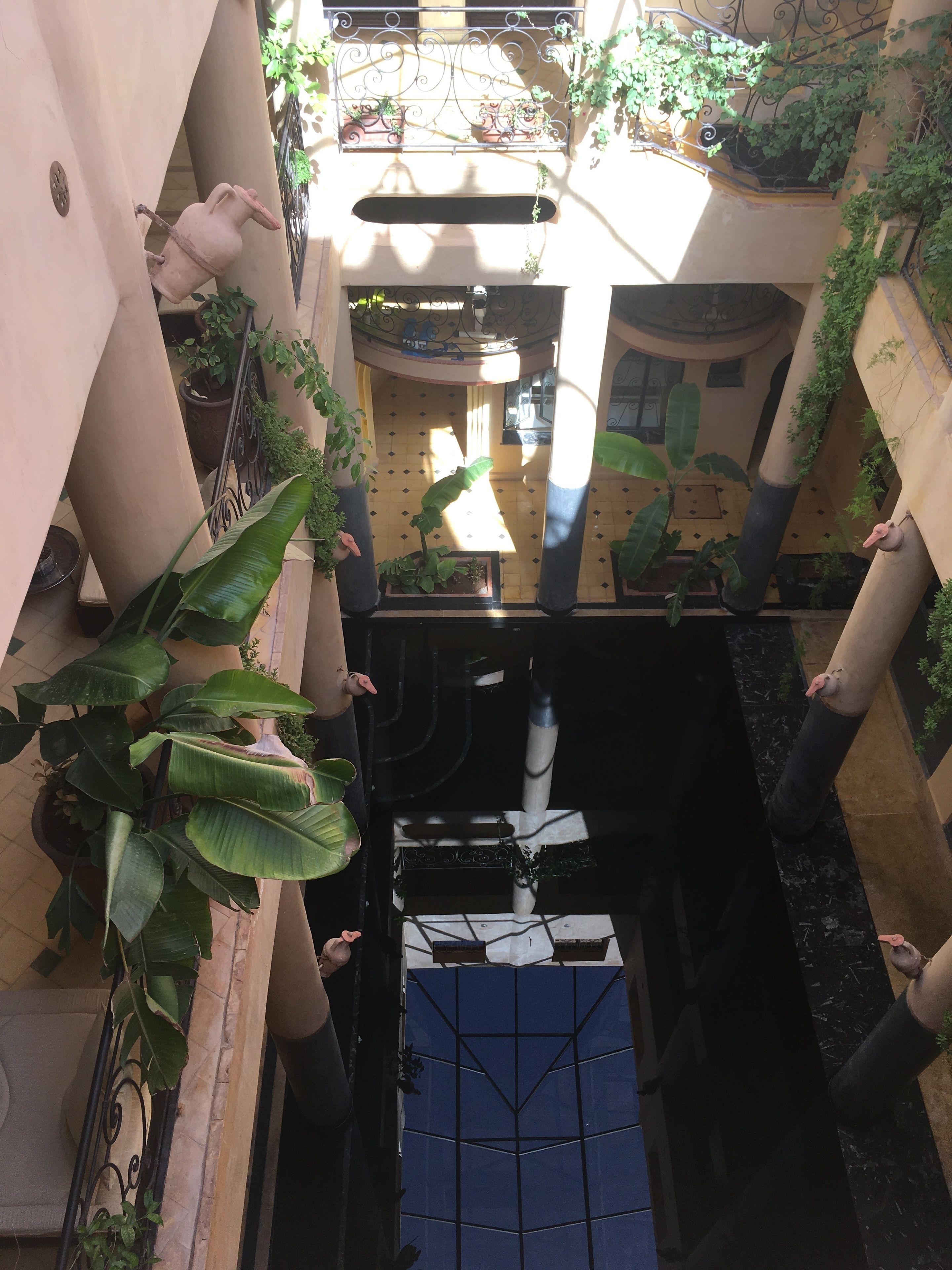 A narrow, enclosed courtyard with a central water feature, surrounded by potted plants and architectural columns.