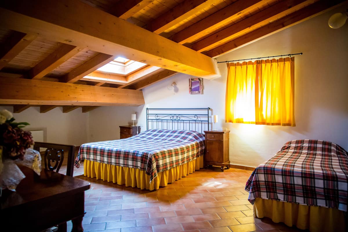 A bedroom with two beds, wooden beams, a skylight, a window with yellow curtains, and a bedside table with a lamp.