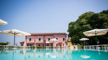 Ein Swimmingpool mit weißen Liegestühlen und Sonnenschirmen, ein rosafarbenes Gebäude mit grünen Fensterläden und ein großer Baum im Hintergrund.