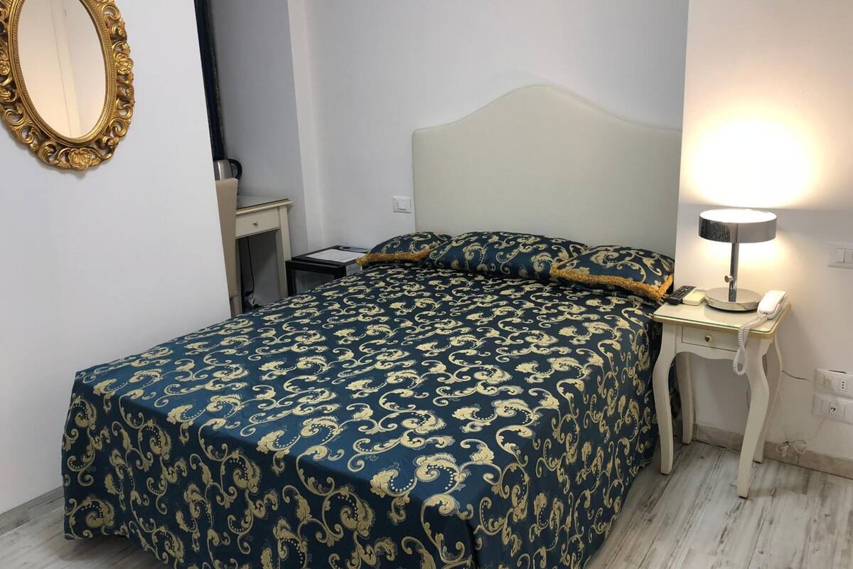 A neatly made bed with a patterned bedspread, a bedside table with a lamp, and a wall-mounted mirror.