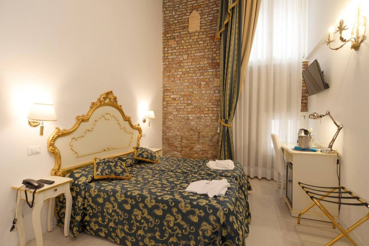 A bedroom with a brick wall, a bed with a gold headboard, a desk with a lamp, and a TV mounted on the wall.