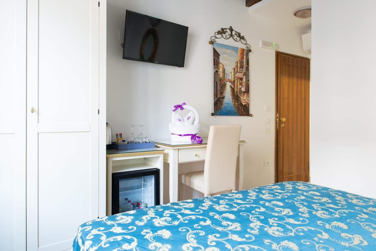 A hotel room with a bed, a desk with a computer, a chair, a small fridge, a TV, and a painting of a bridge.