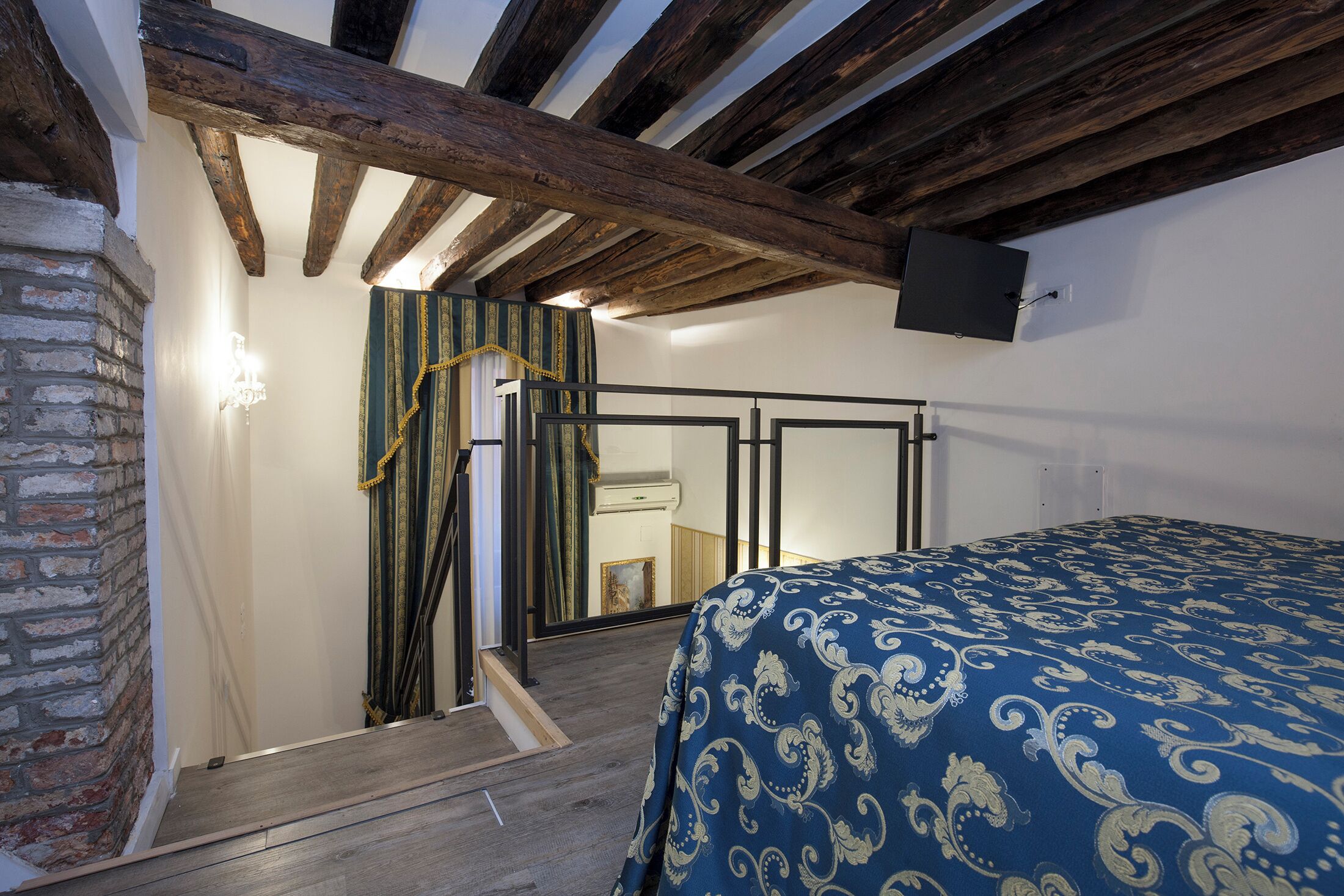 A bedroom with a wooden ceiling, a bed with a patterned bedspread, and a brick wall.
