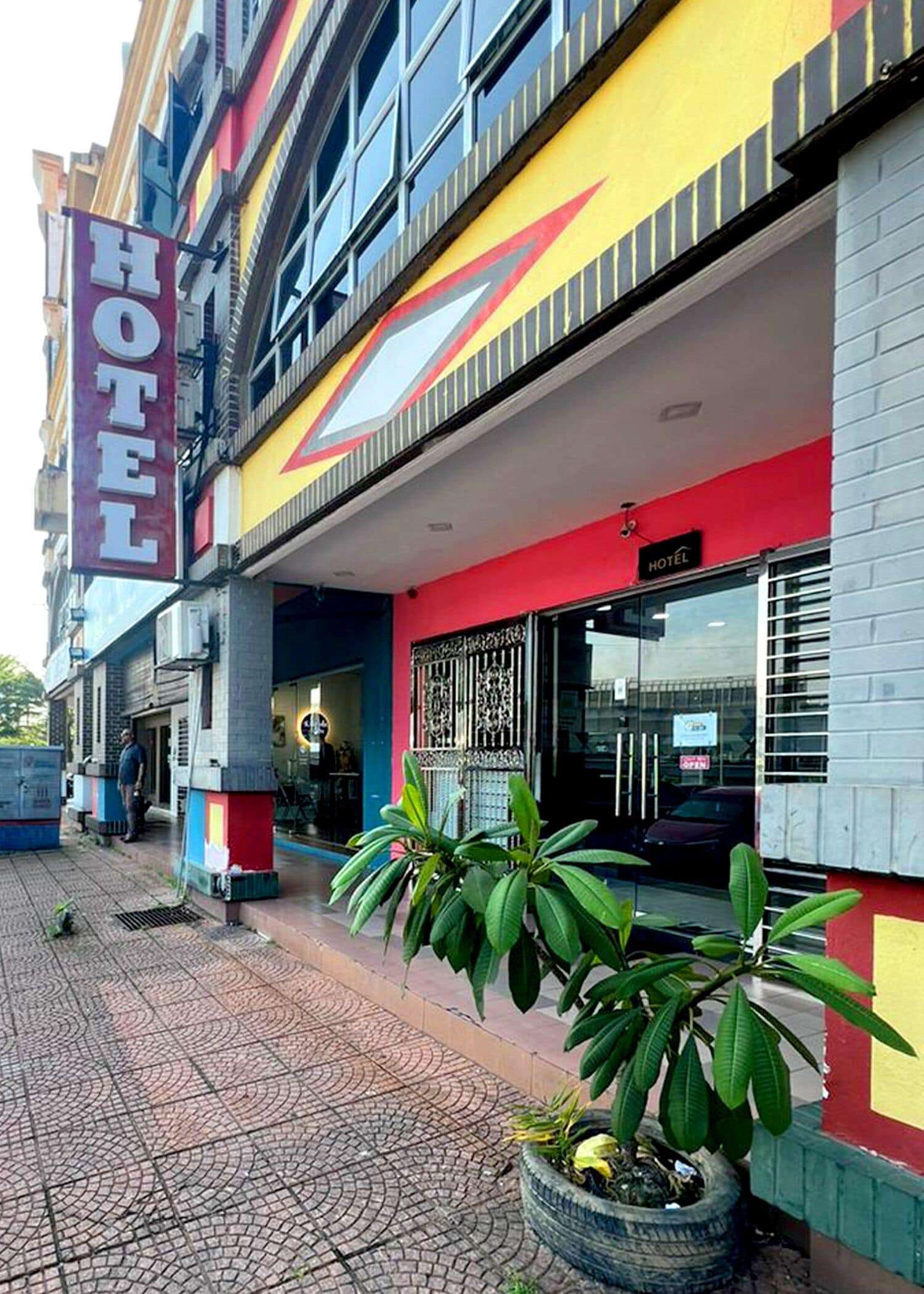 A hotel with a prominent 'HOTEL' sign, a red and yellow facade, and a large potted plant in the foreground.