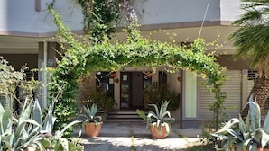 Entrada de um edifício com galeria coberta, vasos de plantas e uma palmeira.