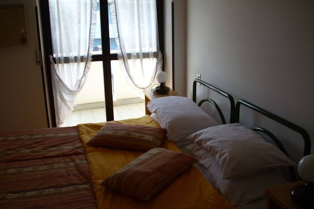 Un dormitorio con cama, almohadas, una mesita de noche y una ventana con cortinas traslúcidas.
