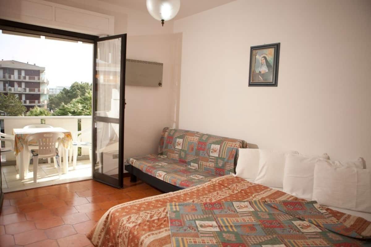 Un dormitorio con cama, sofá, balcón con mesa y sillas, y un cuadro en la pared.