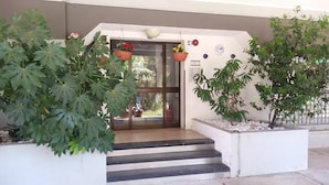 Entrada de edifício com degraus que levam a uma porta, cercada por grandes vasos de plantas e cestos de flores penduradas.