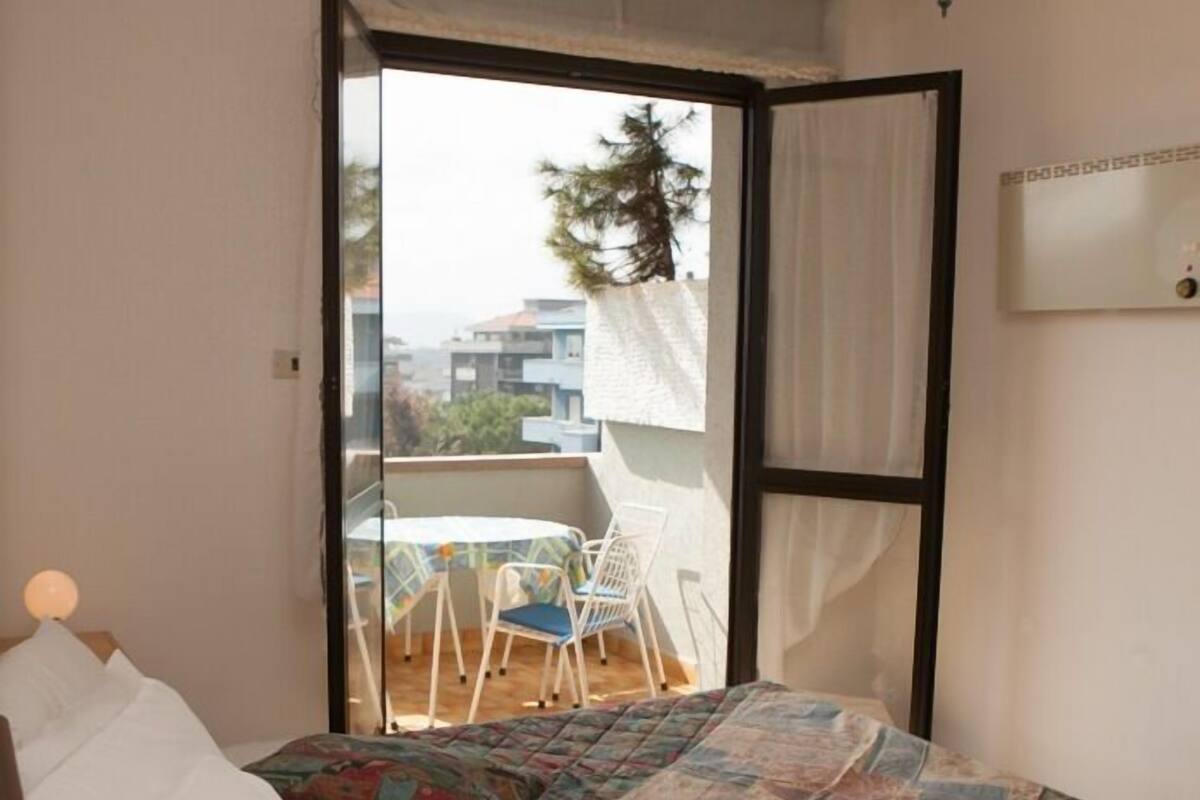 Un dormitorio con cama, mesita de noche con lámpara, ventana con vista a un balcón, y una lámpara colgante.
