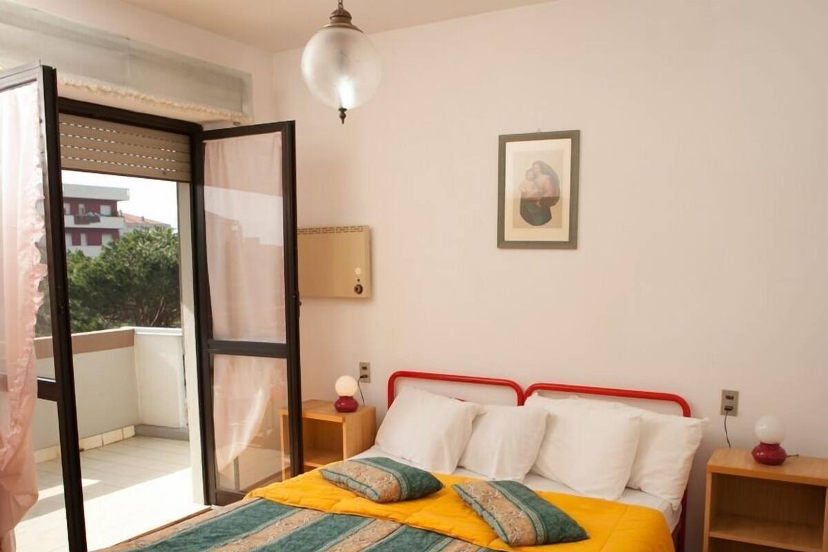 Un dormitorio con cama, mesitas de noche, una ventana con cortinas, una lámpara de pared y un cuadro enmarcado.