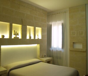 A bedroom with a large bed, a nightstand, a wall-mounted shelf with decorative items, and a window with sheer curtains. - Masseria Don Sante (Fasano)
