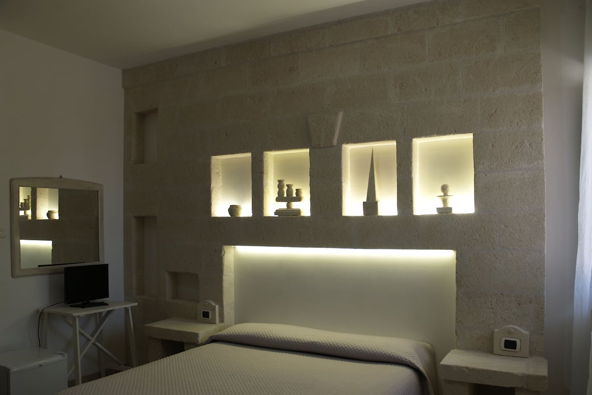 Une chambre d’hôtel avec un grand lit, une table de chevet, un petit bureau avec un ordinateur et une étagère murale agrémentée d’objets décoratifs.