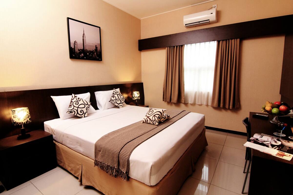 A hotel room with a large bed, a nightstand with a lamp, a wall-mounted air conditioner, a window with curtains, and a table with a fruit bowl and a magazine.