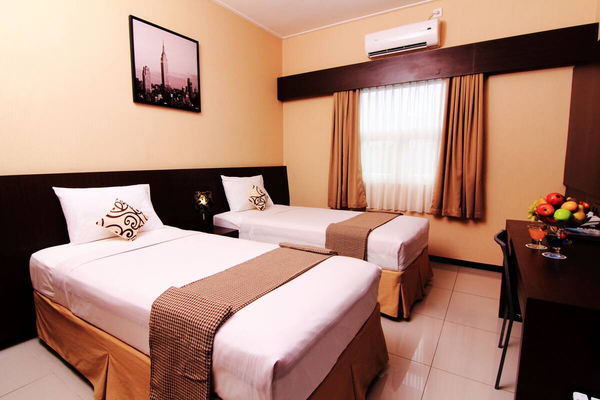 A hotel room with two beds, a desk with a fruit bowl, and a cityscape picture on the wall.