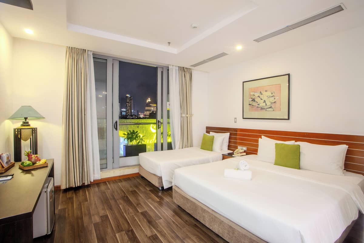 Habitación de hotel con dos camas, un escritorio con lámpara, vista de la ciudad de noche y un cuadro en la pared.