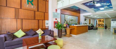 A modern hotel lobby with a reception desk, a sofa with cushions, a coffee table, and a large potted plant.