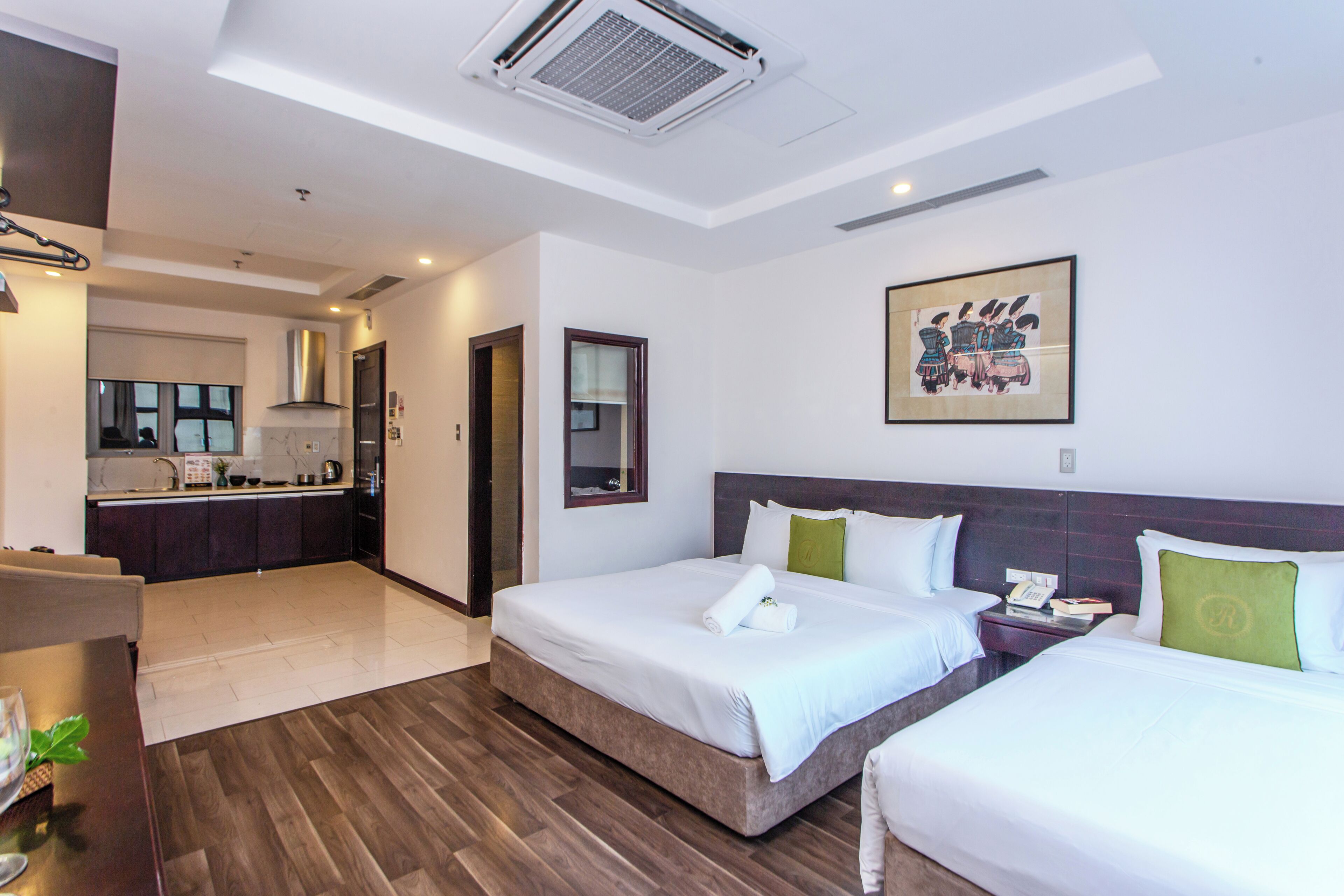 A hotel room with two beds, a small desk, a chair, a dining table, and a kitchen area in the background.